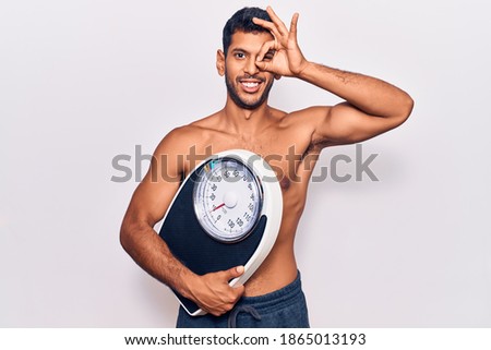 Young latin man shirtless holding weighing machine smiling happy doing ok sign with hand on eye looking through fingers 