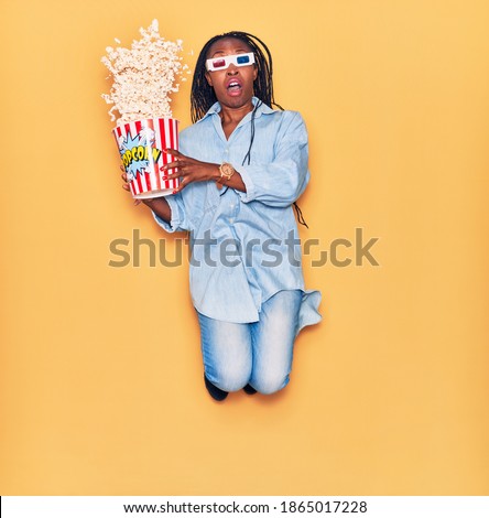 Young beautiful african american woman surprised with open mouth holding bucket of popcorn. Watching movie wearing 3d glasses jumping over isolated yellow background.