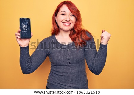 Young beautiful redhead woman holding broken smartphone showing cracked screen screaming proud, celebrating victory and success very excited with raised arm