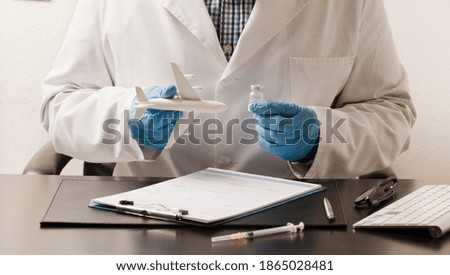 Doctor with blue gloves and airplane in one hand, in the other vaccine the other, desk with syringe, glasses, white form and computer keyboard