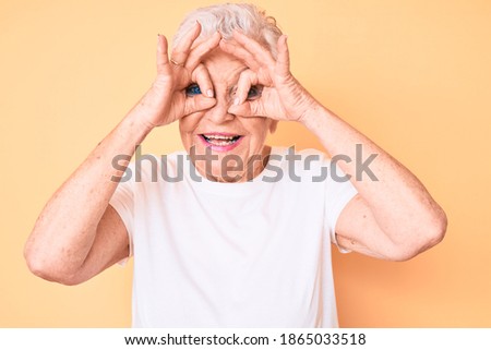 Senior beautiful woman with blue eyes and grey hair wearing classic white tshirt over yellow background doing ok gesture like binoculars sticking tongue out, eyes looking through fingers.