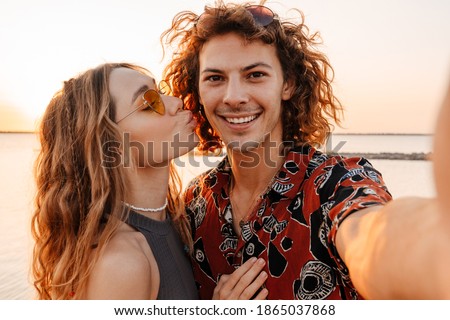 Image of a smiling cheery young loving couple at the beach taking a selfie by camera and kissing