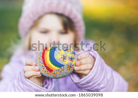 Adorable little toddler girl with painted rainbow on stone during pandemic coronavirus quarantine, outdoors. Child painting rainbow