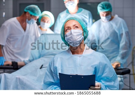 portrait of assistant of doctors making operation to patient, woman in mask and uniform hold digital tablet of patient diagnosis, check, looks up. while colleagues surgeons do operation in background