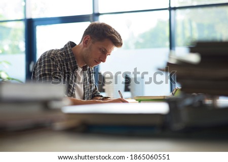 Side view of young male student studying at desk by window in classroom Royalty-Free Stock Photo #1865060551