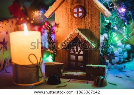A small toy Christmas house with a burning light inside is on a blurred green Christmas tree background. Real estate, holiday, Christmas, miniature. Background for postcards