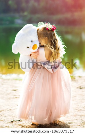 Vertical picture of little blonde girl in pink dress is standing in near the lake holding white teddy bear in her hands at spring sunset time. Tenderness, love and friendship between child and toy.