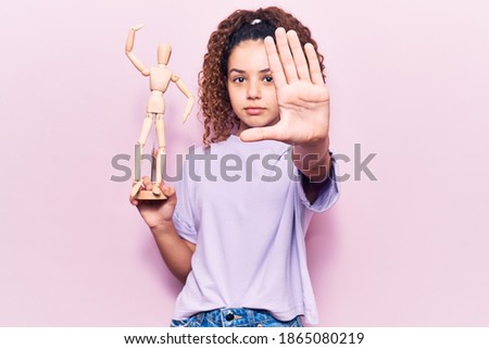 Beautiful kid girl with curly hair holding small wooden manikin with open hand doing stop sign with serious and confident expression, defense gesture 