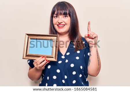 Young plus size woman holding empty frame smiling with an idea or question pointing finger with happy face, number one 