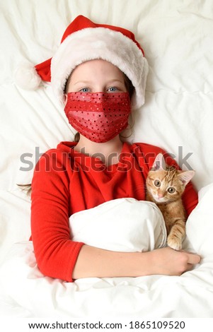 Child girl in the christmas red hat and medical mask hugging her kitten in the bed
