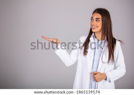 Smiling woman doctor showing aside to copyspace blank place. Confident female medical worker portrait pointing with finger to left side at empty space in studio isolated on gray background