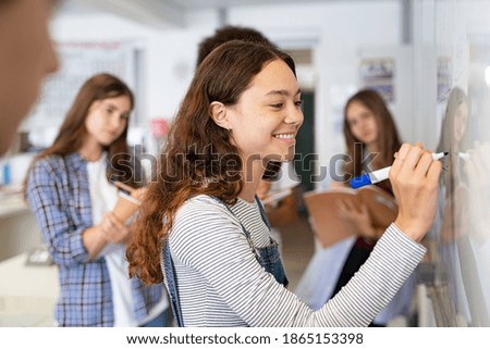 Happy college student writing equation on white board in class. Satisfied young girl solving math problem on whiteboard with classmates in background watching her. Proud high school student writing. Royalty-Free Stock Photo #1865153398