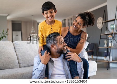 Cheerful african mother and indian father playing with son at home. Cute boy enjoying sitting on father shoulder while looking at camera. Middle eastern family having fun together on the sofa at home. Royalty-Free Stock Photo #1865153404