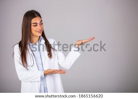 Smiling woman doctor showing aside to copyspace blank place. Confident female medical worker portrait pointing with finger to left side at empty space in studio isolated on gray background