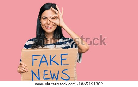 Beautiful young woman holding fake news banner smiling happy doing ok sign with hand on eye looking through fingers 