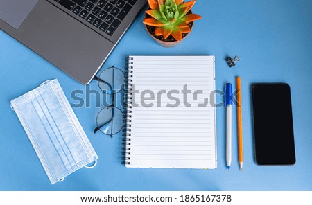 Blue work desk with smartphone, glasses, notepad, pen, medical mask and laptop. Home office and remote work concept. Top view with copy space