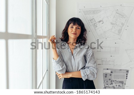 Female architect standing in office beside architecture drawings. Woman entrepreneur standing in office making presentation. Royalty-Free Stock Photo #1865174143