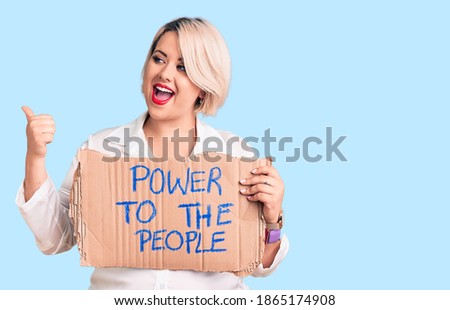 Young blonde plus size woman holding power to the people banner pointing thumb up to the side smiling happy with open mouth 