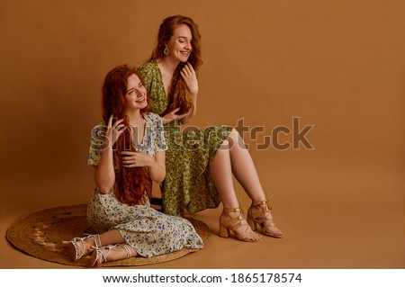 Two beautiful happy smiling women with long natural hair wearing summer floral print dresses, sandals, posing on beige background. Full-length portrait. Fashion, hair care conception. Copy space
