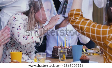 Happy beautiful young Caucasian woman shares birthday fun with friends, blows on cake wearing plastic safety face shield