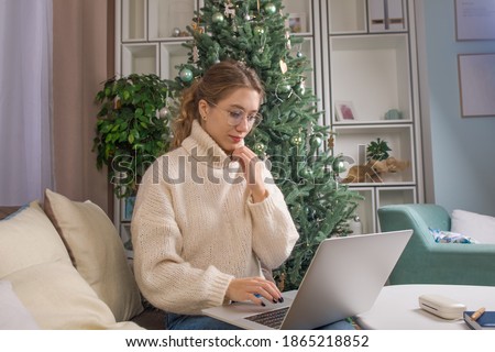 Young female in glasses skilled blogger reading article on website via laptop computer while sitting in home near Christmas tree in winter holidays 