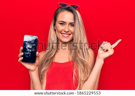 Young beautiful blonde woman holding broken smartphone showing cracked screen smiling happy pointing with hand and finger to the side