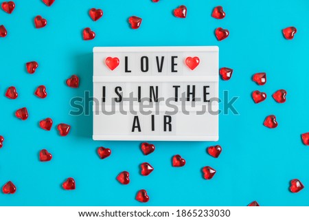Festive banner with glowing lightbox with text Love is in the Air and red glass hearts on blue background for Valentine's Day. Flatlay banner for 14th February. Top view.