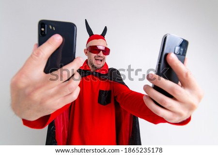 Funny man in black red halloween cape, sunglasses and hat with horns taking selfie with smartphone over white studio background.