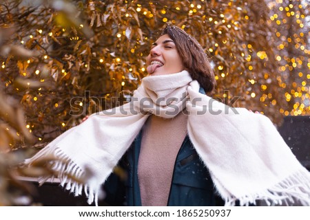 Caucasian happy smiling woman enjoying snow and winter, wearing warm scarf,  christmas lights on background, woman waiting for christmas miracle, street frame 2021