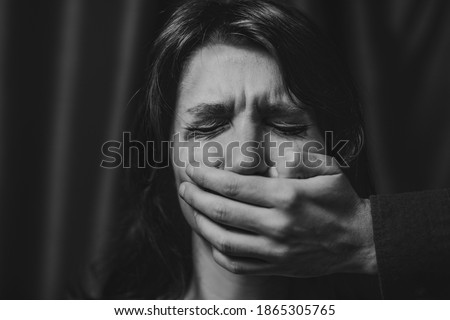 Portrait of a young suffering sad woman with tears in her eyes, a man closes her mouth with his hand. Domestic violence, crying, religion, disagreement, fight, divorce, beating a weaker person, dark. Royalty-Free Stock Photo #1865305765