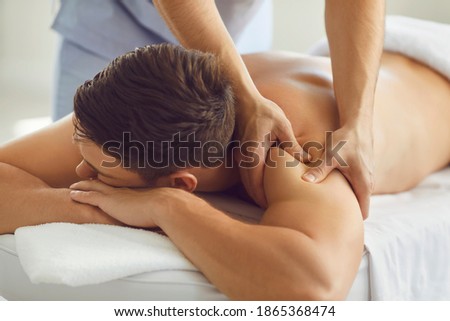 Close-up of relaxed young man lying on massage table enjoying remedial body massage done by professional masseur in modern wellness or health center Royalty-Free Stock Photo #1865368474