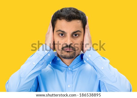 Closeup portrait, handsome young man covering, closing his ears with hands, eyes observing, closed mouth, isolated yellow background. Hear no evil concept. Human emotion, facial expression