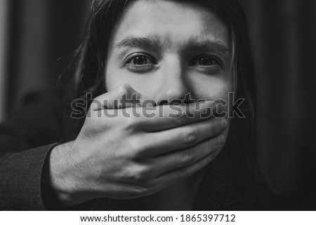 Portrait of a young suffering sad woman with tears in her eyes, a man closes her mouth with his hand. Domestic violence, crying, religion, disagreement, fight, divorce, beating a weaker person, dark. Royalty-Free Stock Photo #1865397712