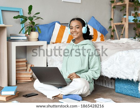 Happy mixed race teen school girl distant college student wearing headphones virtual remote e learning using laptop in bedroom looking at camera. Distance education classes, studying online at home. Royalty-Free Stock Photo #1865428216