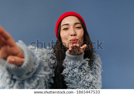 Wonderful stylish girl with brunette long hairstyle in trendy warm cool sweater and red cap blowing kiss and making selfie..
