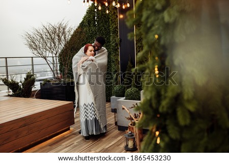 Beautiful couple wrapped in blanket stand at the balcony, handsome African American man hug gorgeous white woman, enjoy tender moments, winter holidays, Christmas morning concept