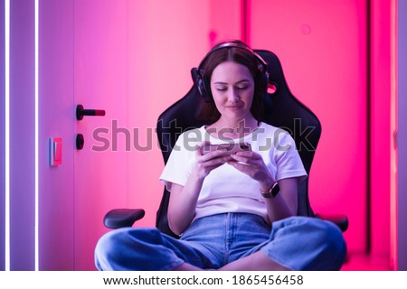 Cybersport gamer playing mobile game on the smart phone sitting on a gaming chair in neon color room. Royalty-Free Stock Photo #1865456458