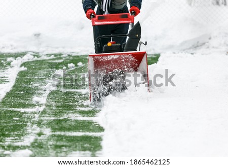 Man using a snow blower to remove large amounts of snow on football field. Man cleans snow with a snow-removing machine on soccer pitch.