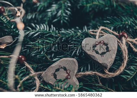 Closeup on christmas composition of wild christmas tree branches and string of wooden hearts with garland print and mixed with fairy lights, blueish green lush color of evergreen and blurry background