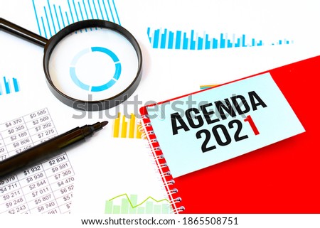 Magnifier,marker and red notebook with white card. Text AGENDA 2021. Financial concept