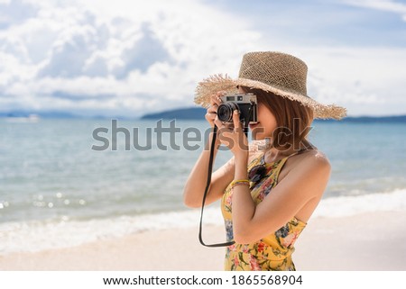Short-haired Asian woman in a yellow floral dress and straw hat standing on the beach take a photo of beautiful seascapes by camera with a blurry sea, mountain and cloudy sky background on a sunny day