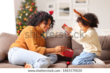 Happy family: ethnic woman sitting on sofa and opening  gifts with boy in Santa hat together while celebrating Christmas at home
