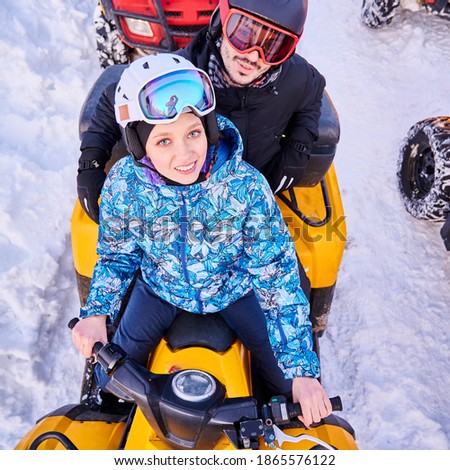 Top view of joyful young woman and man in helmets and goggles driving all-terrain vehicle through snow. Female and male riders looking at camera and smiling while sitting on quad bike.