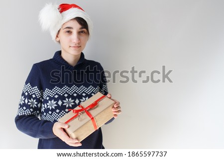 Portrait of a caucasian girl with a short haircut in a New Year's hat and a blue sweater who smiles and holds New Year's gift in her hands on a gray background. Place for text, best view