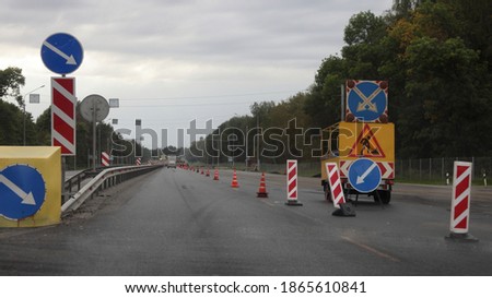 Suburban highway road works, mobile trailer with bright direction signal arrow on blue sign background on right lane of the asphalted road and safety roadsign at summer evening