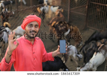 Young indian farmer showing smart phone at goat dairy farm