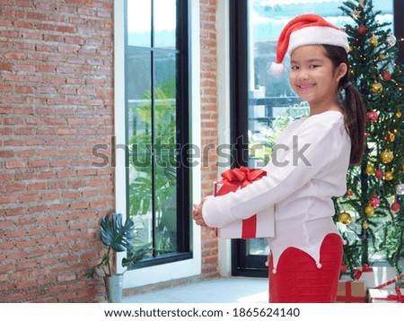 Little girl in santa hat holding gift box at home. Merry Christmas, Happy New Year.