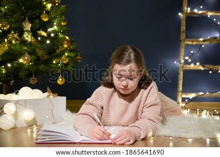 Cute little child girl writes the letter to Santa Claus near Christmas tree indoors.