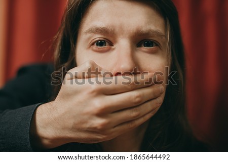 Portrait of a young suffering sad woman with tears in her eyes, a man closes her mouth with his hand. Domestic violence, crying, religion, disagreement, fight, divorce, beating a weaker person, dark. Royalty-Free Stock Photo #1865644492