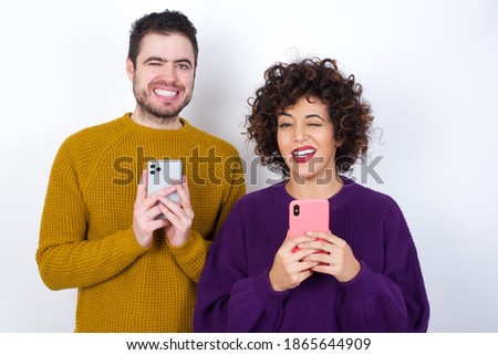 Pleased Young couple wearing knitted sweater standing against white background using self phone and looking and winking at the camera. Flirt and coquettish concept.